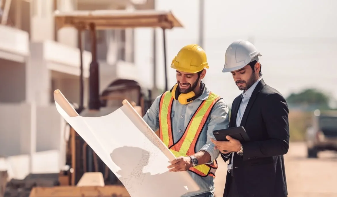 Two project managers at a construction site reviewing blueprints and using a tablet for project coordination and contractor management.